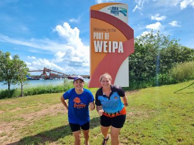 Two women standing in the running position in front of a sign that says Port of Weipa with the port in the background.